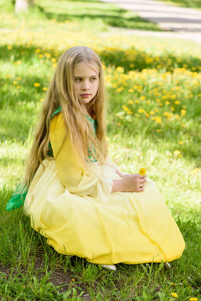 Blond young girl posing in a yellow green dress sitting on the grass with dandelions yellow flowers - Foto, Imagem