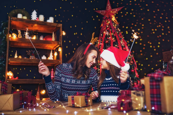holidays, family and people concept. Happy mother and little girl in santa helper hat with sparklers in hands, gift boxes on foreground Having fun - Fotoğraf, Görsel
