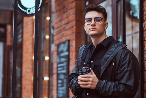 Молодой красивый мужчина держит кофе на вынос, стоя возле кафе на улице
. - Фото, изображение
