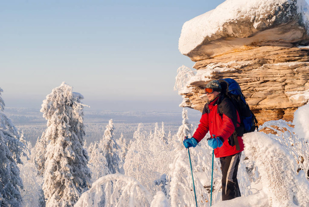 смеющийся путешественник с рюкзаком и палками для нордической ходьбы стоя в морозном зимнем пейзаже рядом с красивой скалой и смотрит вдаль
 - Фото, изображение