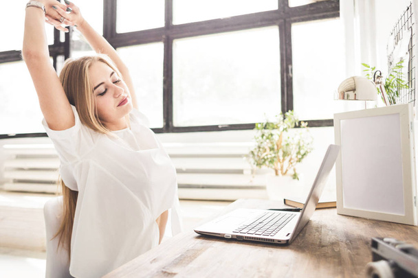 Young business woman working at home behind a laptop and stretching her hands. Creative Scandinavian style workspace - Фото, изображение
