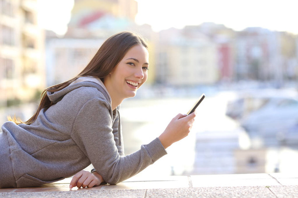 Feliz adolescente sosteniendo un teléfono inteligente mirando a la cámara acostada en la acera de una ciudad costera
 - Foto, imagen