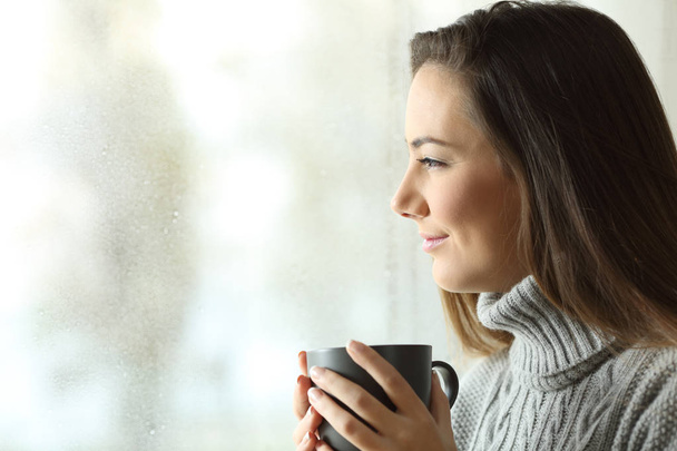 Happy woman looking at rain holding coffee through the window of home or hotel - Fotoğraf, Görsel