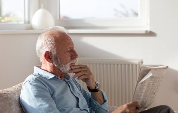Vanhempi mies istuu sohvalla olohuoneessa ja lukee sanomalehteä
 - Valokuva, kuva