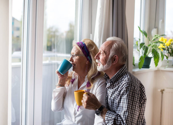 Happy senior couple standing beside window and drinking tea - Zdjęcie, obraz