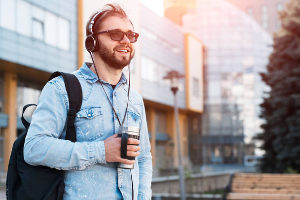 Modern mosolygó szakállas csípő a fekete hátizsák, fejhallgató, a fül és a thermo bögre kávét a kezében - Fotó, kép