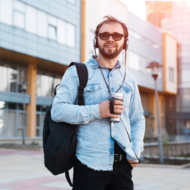 Modern smiling bearded hipster with black backpack, headphones in ears and thermo mug with coffee in hand - Φωτογραφία, εικόνα