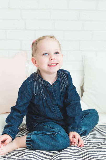 Cute little girl in blue denim overalls sitting on the bed. - 写真・画像