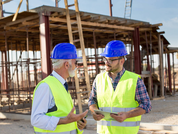 Kaksi tyytyväistä insinööriä, joilla on tabletti, puhuvat rakennustyömaalla
 - Valokuva, kuva