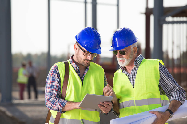 Twee tevreden ingenieurs kijken blauwdrukken op de bouwplaats met metalen constructie in achtergrond  - Foto, afbeelding
