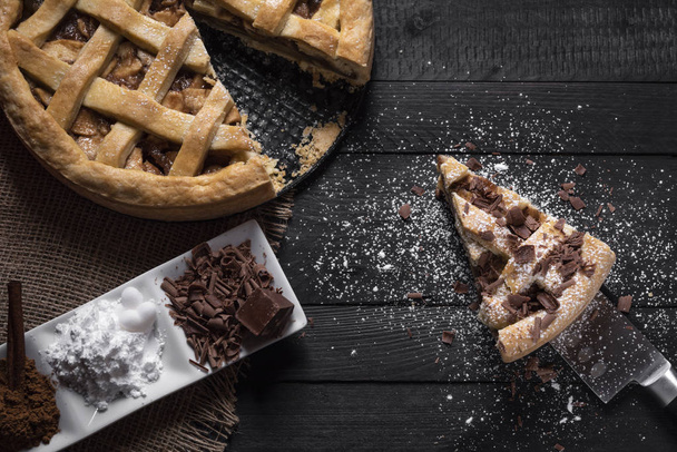 Kitchen table with a whole apple pie and one slice on a knife, with powdered sugar and chocolate and a plate with sweet seasoning. Above view. - Photo, Image