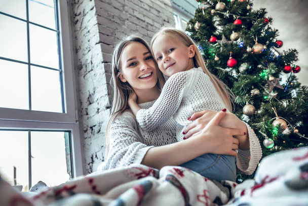Cheerful mother and daughter sitting on the bed near beautiful decorated Christmas tree, spending New Year eve at home together, happy family concept - Foto, afbeelding