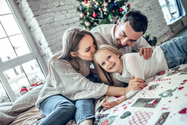A family of three hugging in front of the christmas tree on the bed. New year's eve. Cozy holiday at the fir-tree. love, happiness and big family concept - Zdjęcie, obraz