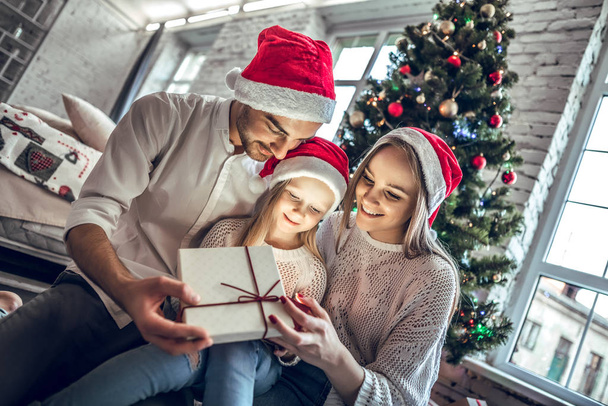Young mother, father and little daughters opening a magical Christmas gift by a Christmas tree in cozy living room in winter - Valokuva, kuva