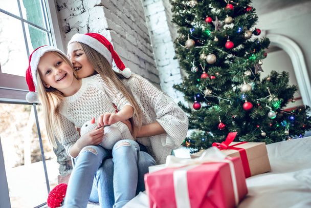 Young mother and cute girl having fun on christmas eve in living room. Xmas holiday lifestyle. Beautiful christmas tree and white brick wall in the background. - Foto, Imagen