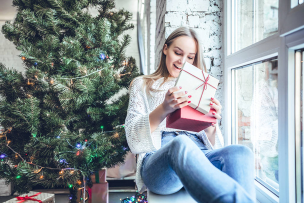 A beautiful young woman in a white sweater dress unpacks New Year's presents near New Year tree. Front view - Фото, изображение
