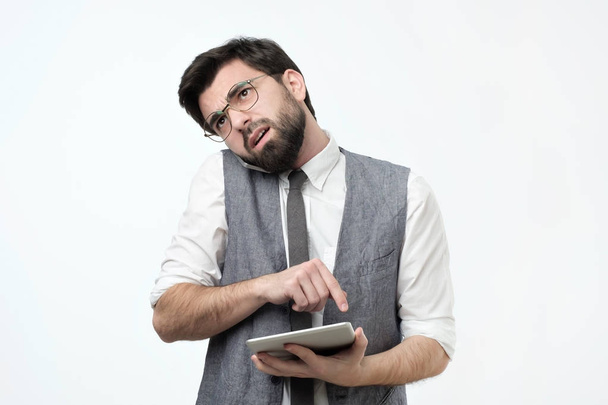 verwirrter junger hispanischer Typ mit Bart, der auf einem Mobiltelefon auf weißem Hintergrund spricht. Er versucht, die beste Lösung aus Finanzproblemen zu finden. ich höre dir aufmerksam zu. - Foto, Bild
