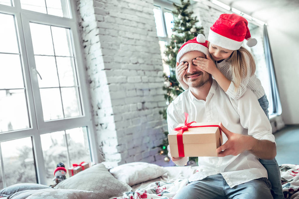 Cute little girl is giving her handsome father a gift box. Both are sitting on bed near X-mas tree at home and smiling - Фото, зображення