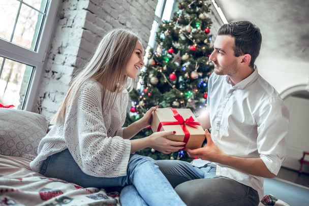 Beautiful young couple is celebrating at home. Pretty girl is giving her boyfriend a gift box - 写真・画像