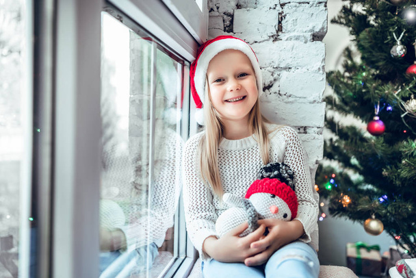 A little child sits on a window sill. Concept Happy Christmas, New Year, holiday, winter, childhood. - Foto, afbeelding