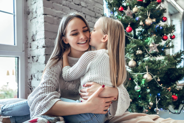 Sweet little girl is hugging and kissing her beautiful young mom in cheek while sitting on bed at home on x-mas tree background - Foto, Imagem