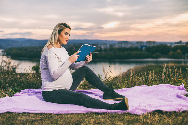 Beautiful pregnant woman enjoys reading book outdoor.Toned image. - 写真・画像