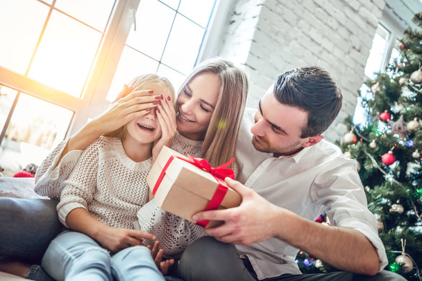 Portrait of young happy family. mother covers his daughter s eyes as father going to make surprise for daughter gives present, near Christmas tree. Happy smiling girl receives gift from dad. Surprise concept - Photo, Image