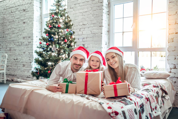 Family in Christmas Santa hats lying on bed. Mother father and baby having fun in bedroom. People relaxing at home. Winter holiday Xmas and New Year concept - Photo, image