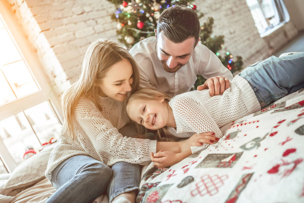 Happy family portrait of mother and girl child laying on cosy bed in festively decorated room with Christmas tree. New Year s celebration. - Photo, image