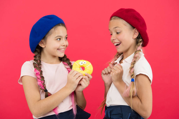 Kids huge fans of baked donuts. Share sweet donut. Girls in beret hats hold glazed donut red background. Kids playful girls ready eat donut. Friendship and generosity. Sweets shop and bakery concept - Fotó, kép