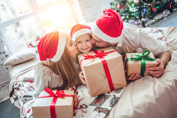 Christmas. Family. Happiness. Parents are kissing her little daughter in Santa hats while sitting on a bed at home near the Christmas tree - Фото, изображение