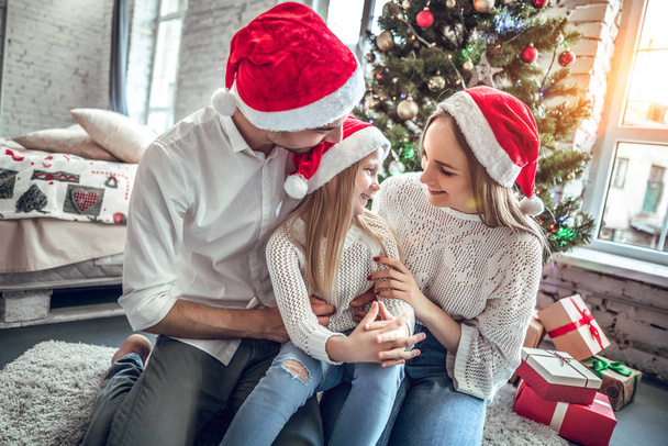 People, family, celebration and holidays concept. Mother, father and daughter with attractive appearance look at each other s eyes, sit on white carpet near decorated New Year tree and gift box - Foto, Imagen