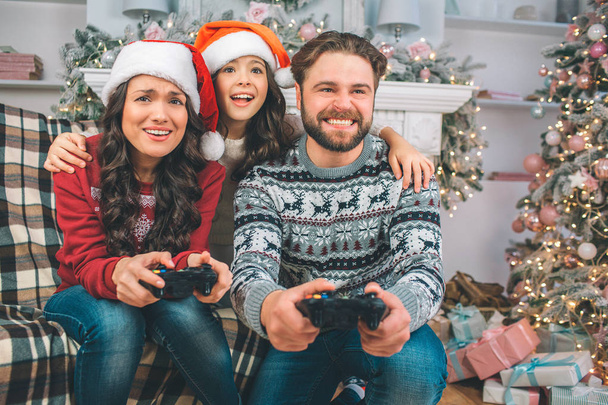 Playful and happy family sitting on sofa and playing games using joystick. Child is between parents. Young man and woman playing games. They look festive. - 写真・画像