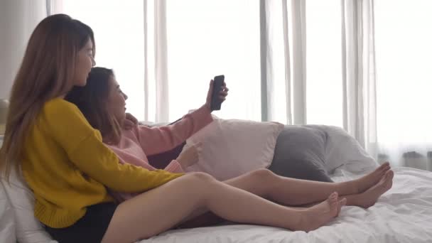 Beautiful young asian women LGBT lesbian happy couple sitting on bed hug and using phone taking selfie together bedroom at home. LGBT lesbian couple together indoors concept. Spending nice time home. - Footage, Video