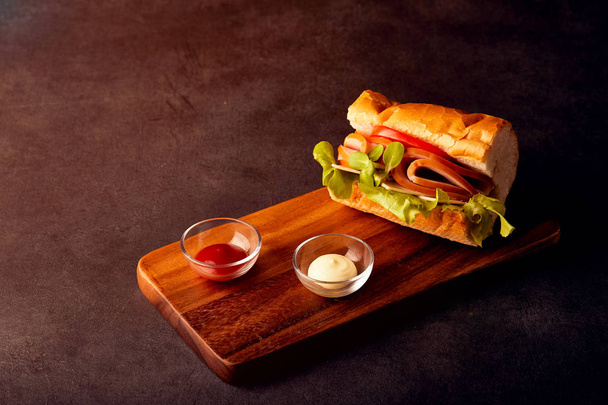 Sanduíche delicioso com presunto, tomate, arugula, juntamente com molho de tomate e maionese colocação em placa de madeira em luz escura
 - Foto, Imagem