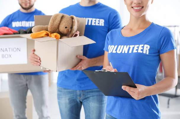 Молодая женщина-волонтер с планшетом и её командой держит коробки с пожертвованиями в помещении
 - Фото, изображение