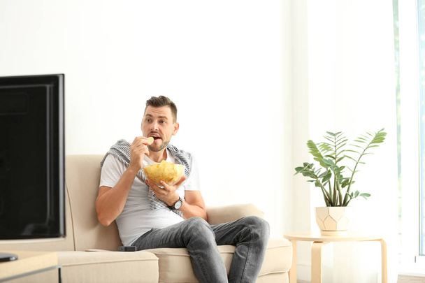 Man with bowl of potato chips watching TV on sofa in living room - Foto, Bild