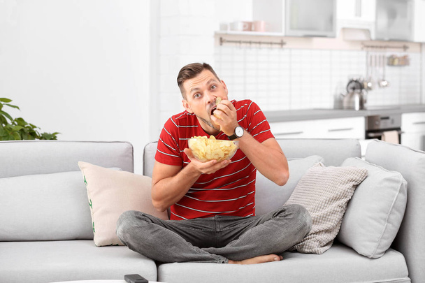 Mann mit Schüssel Kartoffelchips sitzt auf Sofa im Wohnzimmer - Foto, Bild