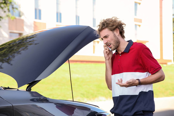 Молодой человек разговаривает по телефону рядом со сломанной машиной, на улице
 - Фото, изображение