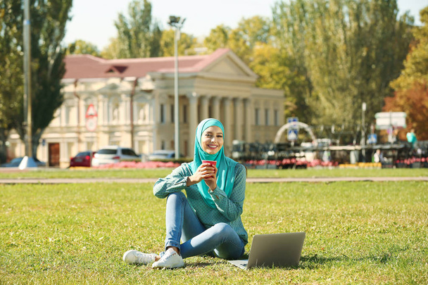 Muslim woman with cup of coffee and laptop sitting on green lawn outdoors - Fotografie, Obrázek