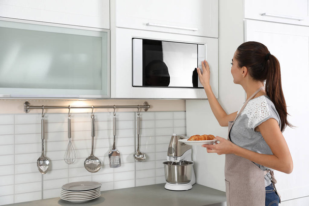 Young woman with plate of croissants near microwave oven in kitchen - Photo, image