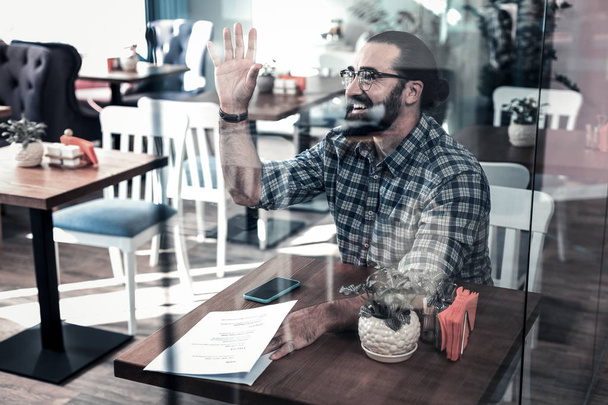 Pleasant bearded man smiling and waving his hand meeting friend in cafeteria - Фото, изображение