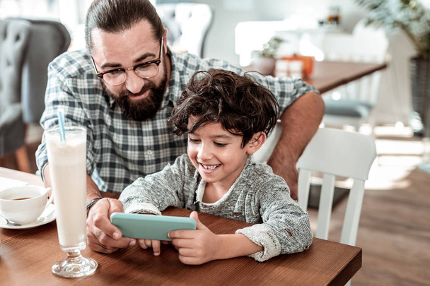 Playing games. Beaming good-looking father and son feeling entertained playing games on the smartphone - Foto, imagen