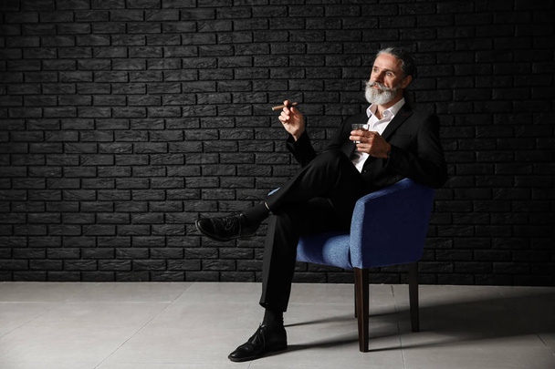 Ανώτερος άνθρωπος πίνει ουίσκι και πούρα κάπνισμα κοντά σκοτεινό τούβλο τοίχο - Φωτογραφία, εικόνα