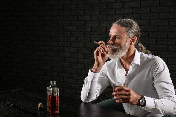 Старший чоловік п'є віскі і курить сигару на темному тлі
 - Фото, зображення