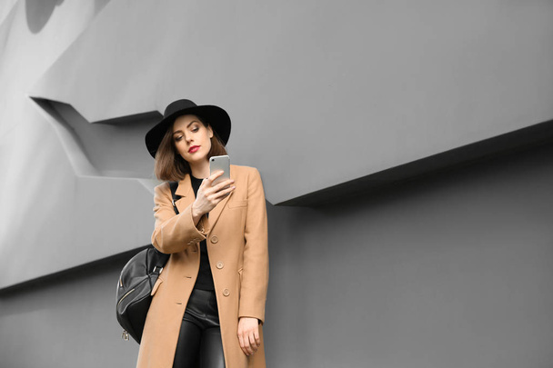 Красива модна жінка з мобільним телефоном біля сірої стіни на відкритому повітрі
 - Фото, зображення