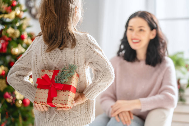 Merry Christmas and Happy Holidays! Cheerful mom and her cute daughter girl exchanging gifts. Parent and little child having fun near tree indoors. Loving family with presents in room. - Zdjęcie, obraz