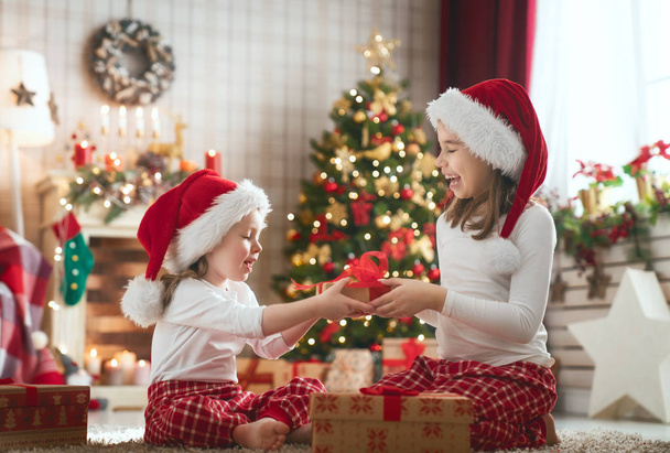 Merry Christmas and Happy Holidays! Cheerful cute childrens girls opening gifts. Kids wearing pajamas having fun near tree in the morning. Loving family with presents in room. - Фото, изображение
