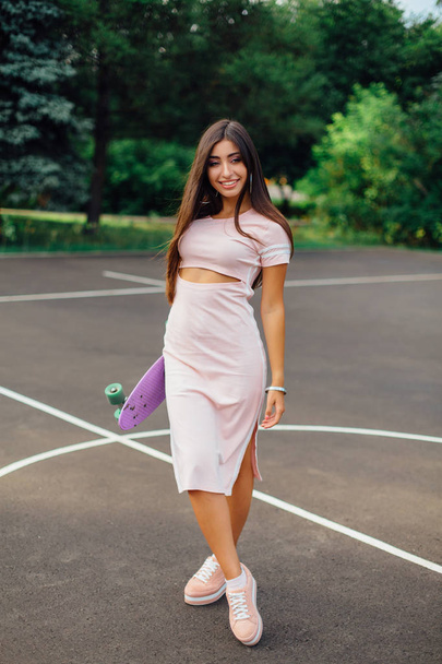 Portrait of a smiling charming brunette female holding in hand her skateboard on a basketball court. Happy woman with trendy look taking break during sunset. - Foto, Imagem