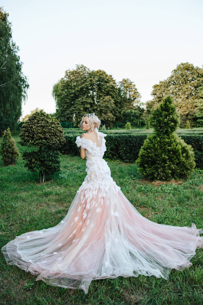 blonde woman in creative wedding dress with feathers. outdoor portrait.  caucasian model in city park. lady in white evening dress. trendy messy hairstyle. romantic concept - Foto, Imagen